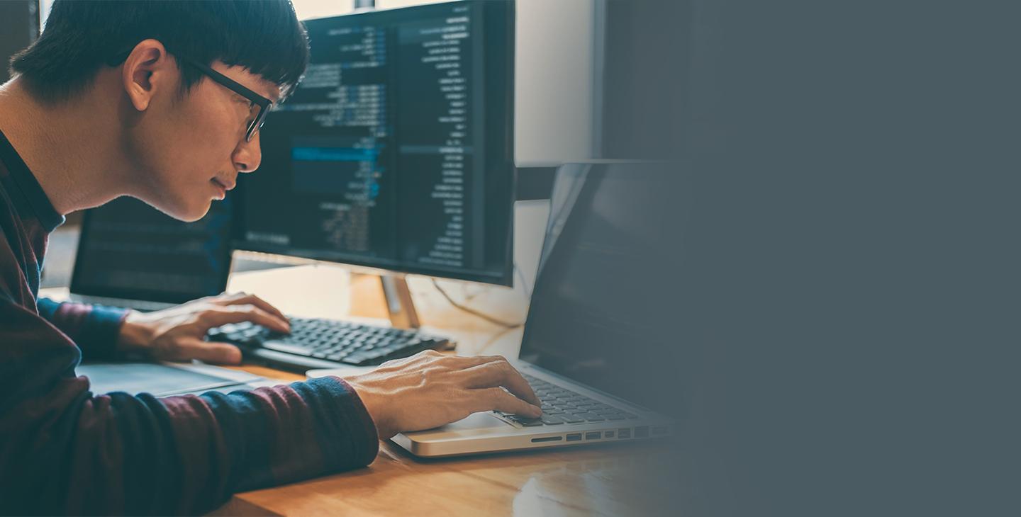 Man wearing glasses and a blue and maroon striped shirt working on cybersecurity protection on a laptop and desktop computer simultaneously